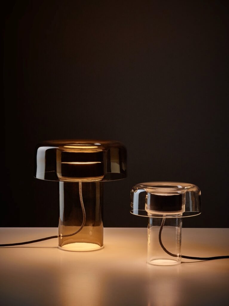 Brokis Spectra Table lamps with sculptural glass bases cast a soft, golden glow on a white surface against a dark backdrop.