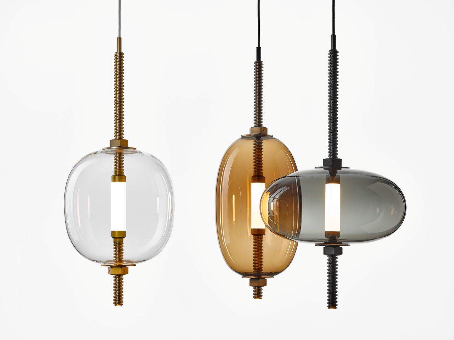 Three Brokis Under Pressure pendants with handblown glass shades - clear, brown, grey - glow against a crisp white backdrop.