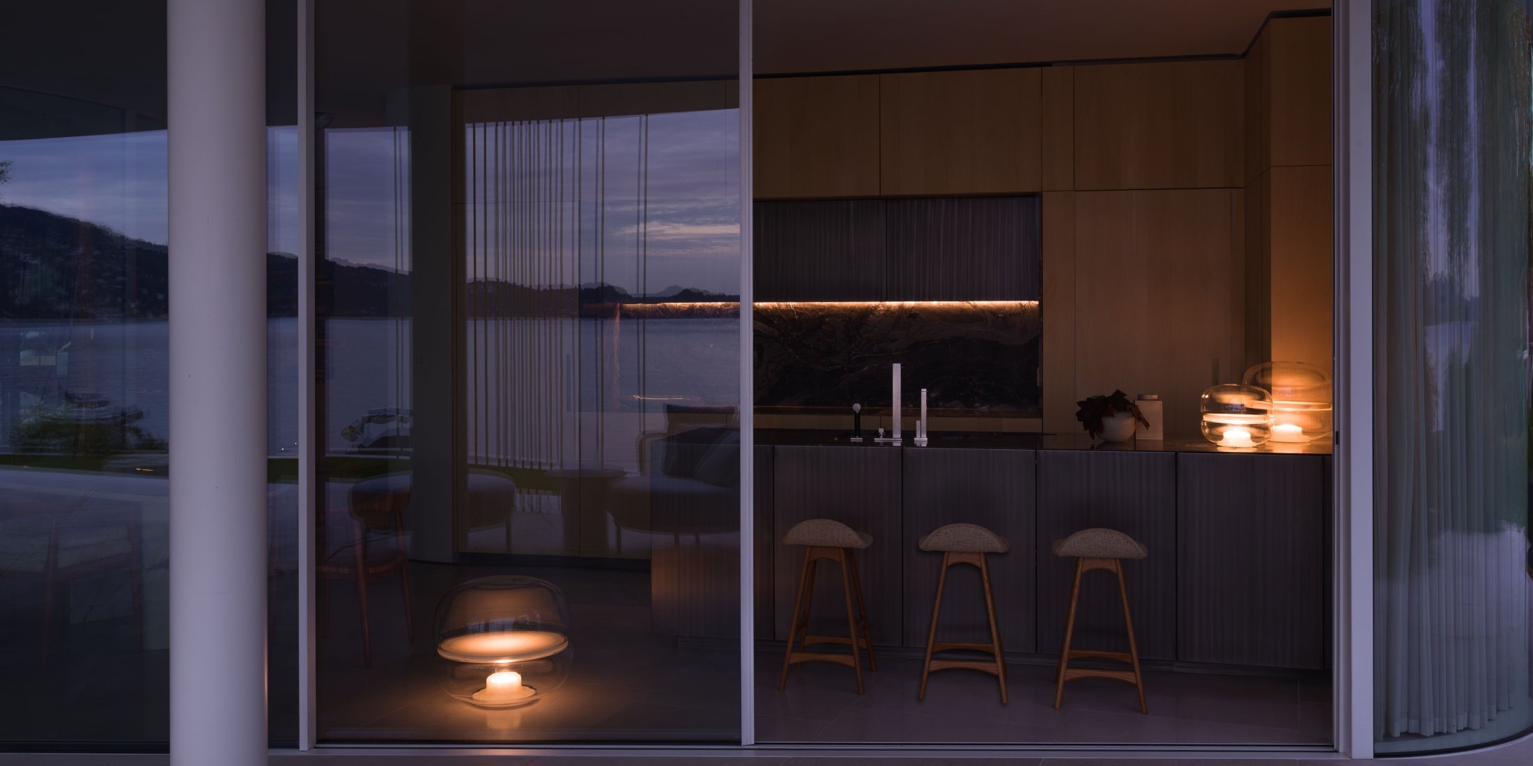 Brokis Macaron lamps in a modern kitchen glows at dusk, with warm wood, overlooking a tranquil lake and hills.