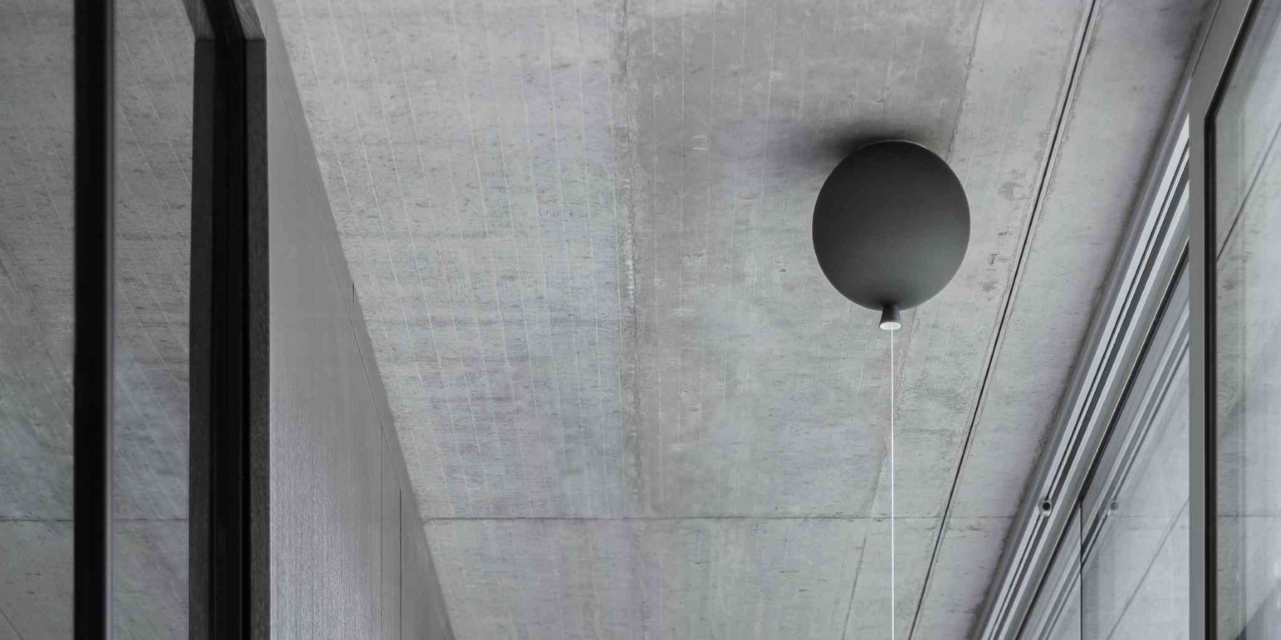 Black Brokis Memory ceiling light with visible flex hangs from concrete amid reflective glass and metal walls.