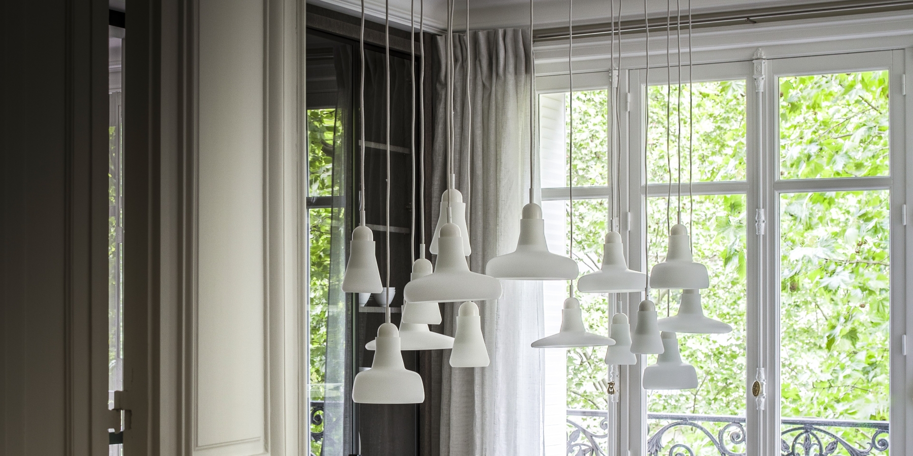 Brokis Shadows white pendants hang before tall windows draped in pale curtains, bathed in natural light and leafy views.