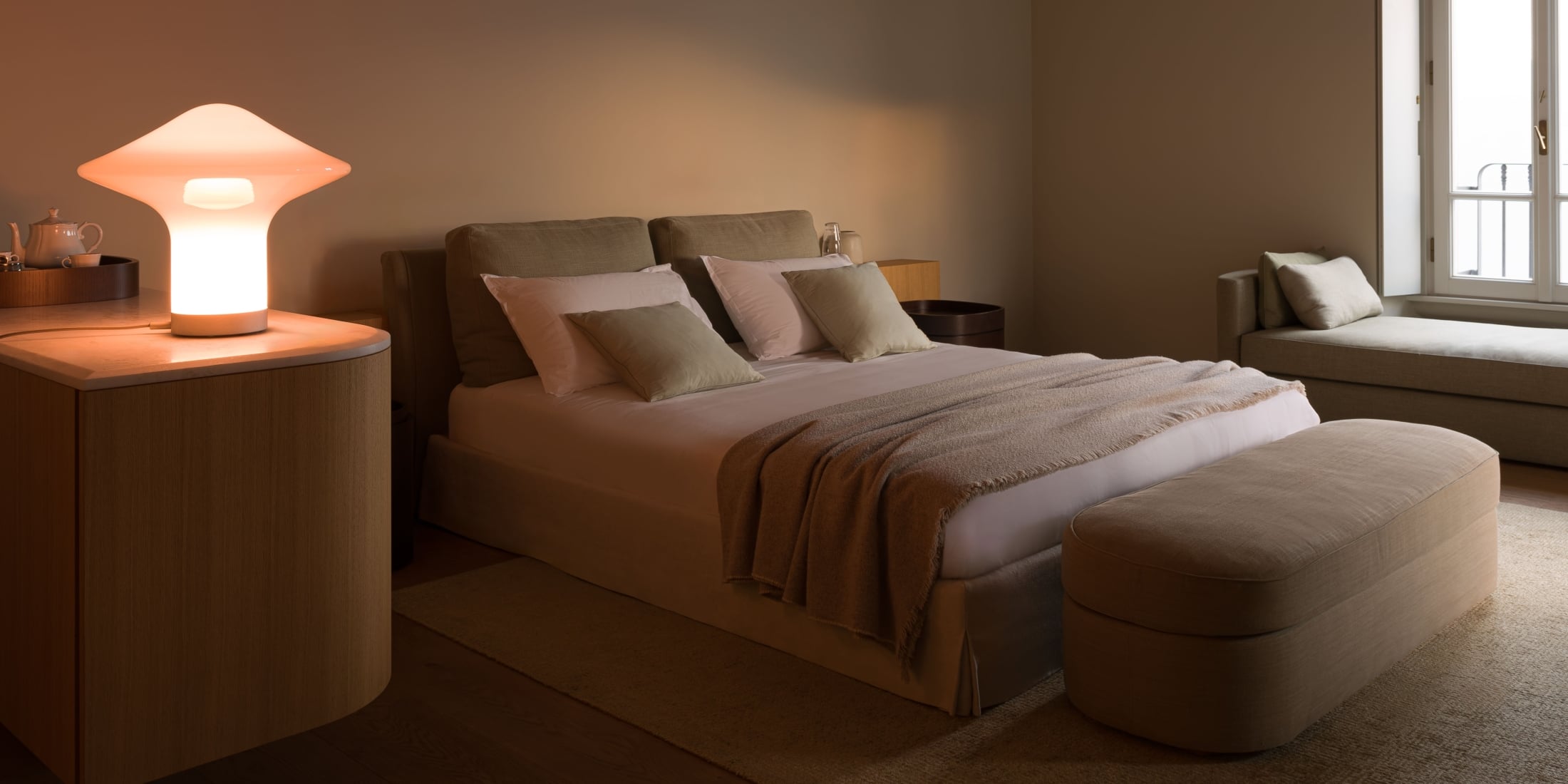 Brokis Trottola Table L atop a wooden bedside table in a luxe bedroom with a beige bed, and plush pillows.