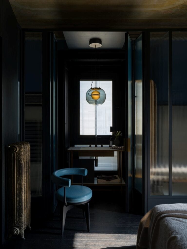 A moody modern bathroom with a frosted glass door, blue chair, Brokis Big One Large pendant, and minimalist dark décor.