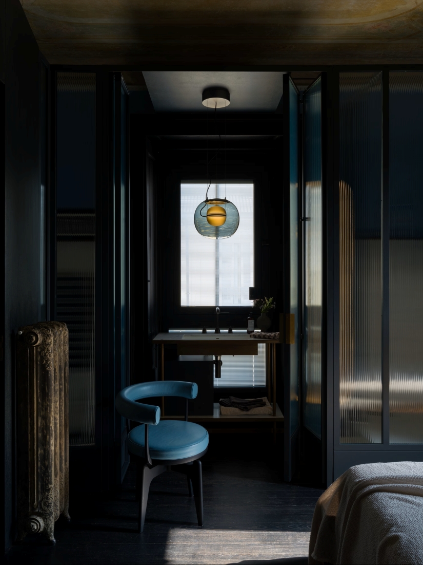 A moody modern bathroom with a frosted glass door, blue chair, Brokis Big One Large pendant, and minimalist dark décor.