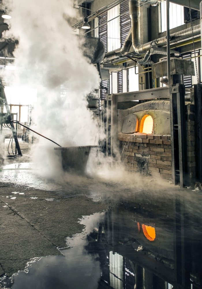 A Brokis furnace casts a warm glow in an industrial factory; steam rises, reflected on the floor amid pipes and windows.