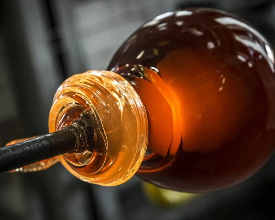 Brokis molten glass, glowing amber and orange, is artfully shaped on a metal pipe amid a soft grey blur.