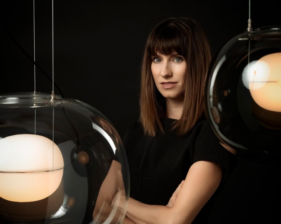 Lucie Koldolva stands arms folded, framed by Brokis Big One pendants glowing elegantly.