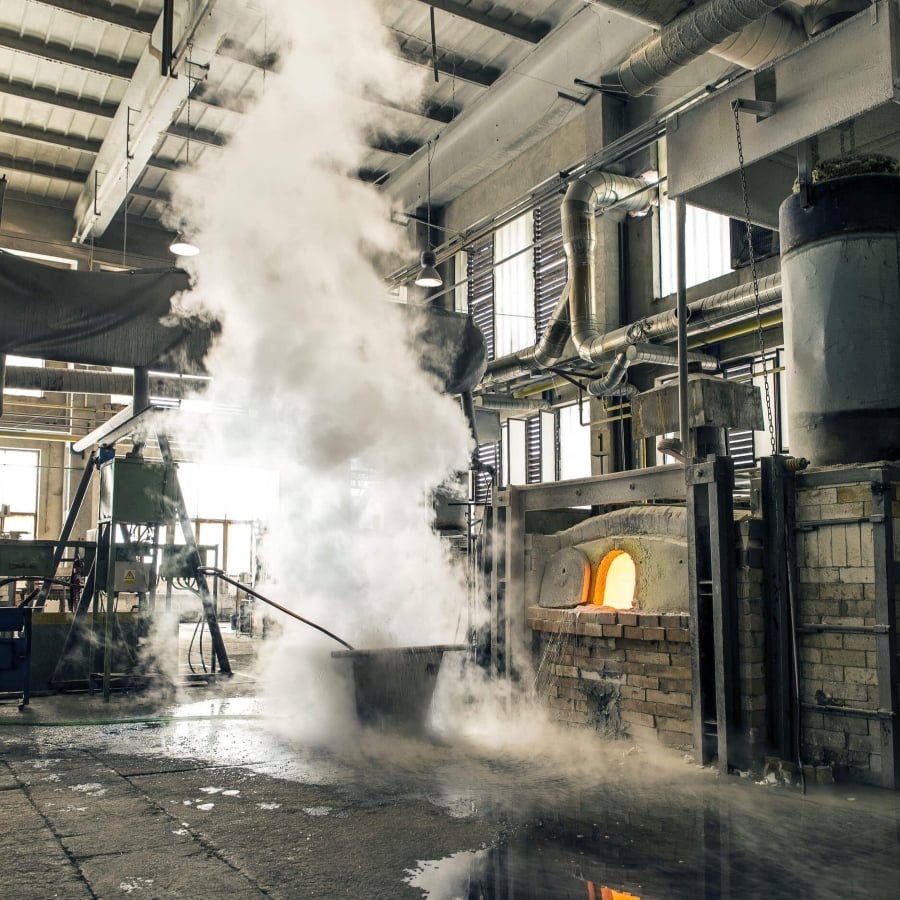 A glass furnace glows in a Brokis factory, steam swirling past exposed pipes and reflections on polished concrete.