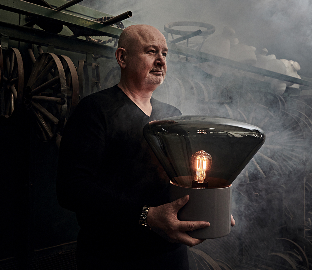 Jan Rabell holds a Brokis Muffins lamp in a smoky, industrial space with looming metal machinery.