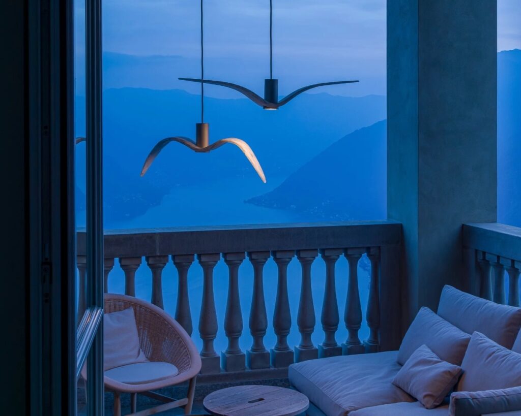 A luxurious dusk balcony with a cushioned sofa, wicker chair, and Brokis Night Bird lights overlooks serene blue mountains.