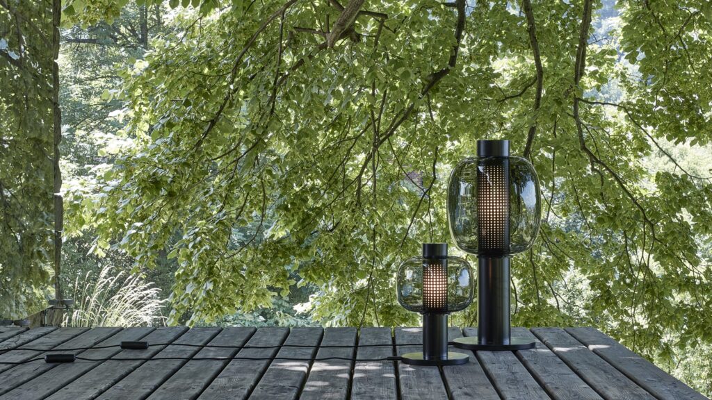 Sustainable Brokis lanterns with black bases grace a weathered deck amid sunlit foliage, exuding refined serenity.