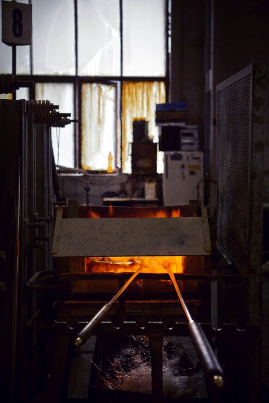 Two metal rods glow in a furnace, their fiery radiance illuminating the Brokis glassworks and a large window beyond.