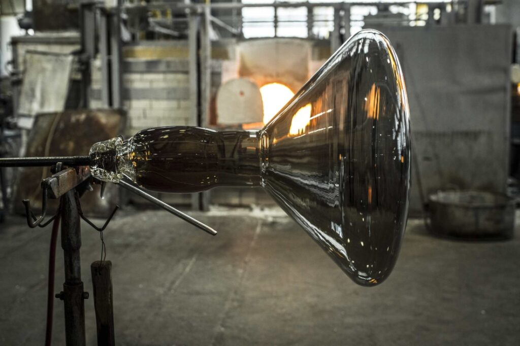 A glassmaker shapes molten glass at the end of a blowpipe; a Brokis furnace glows amid metal tools in the workshop.