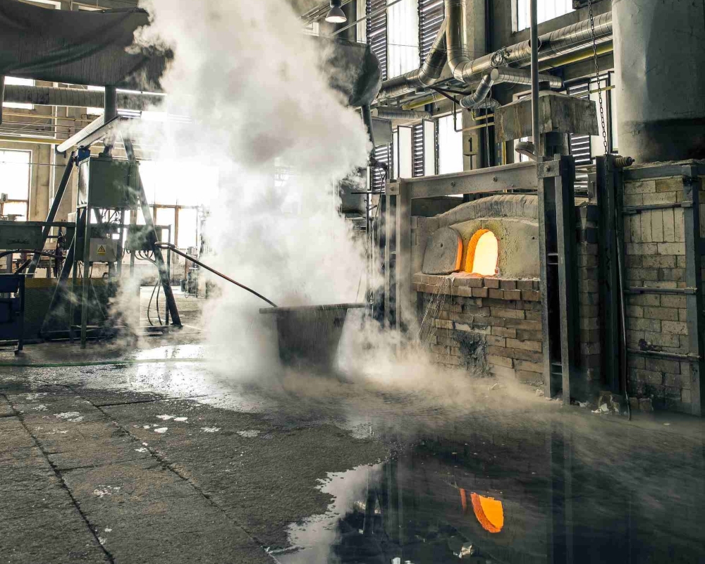 A brick furnace radiates orange light amid industrial pipes and steam, reflecting on the wet floor of a Brokis factory.