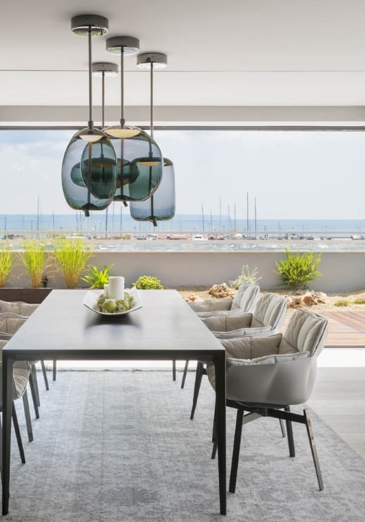 A modern dining room with a black table, cushioned chairs, and Brokis Knot pendants above; ocean views beyond.