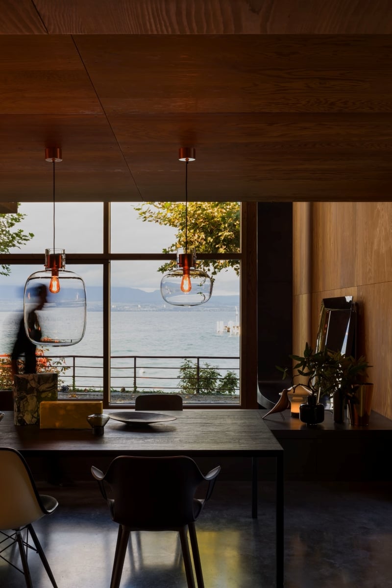 Brokis Awa pendants in a modern dining room with wood walls, black chairs, and lake views framed by large windows.
