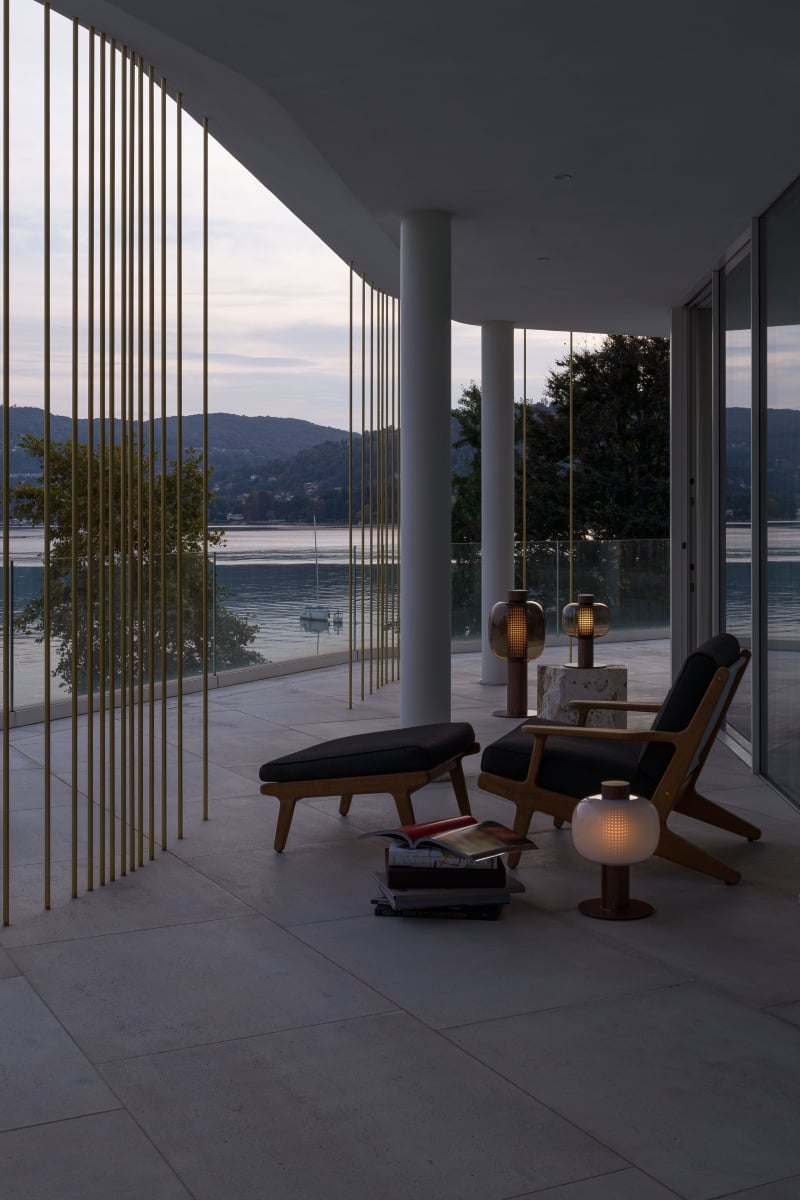 Brokis Bonbori lamps glowing by elegant lounge chairs, gold rails, and glass walls, at a modern lakeside terrace at dusk.