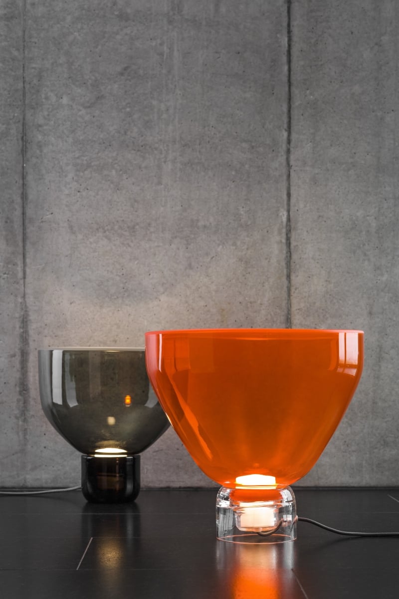 Brokis Lightline table lamps - one vivid orange, one smoke grey - glow on a dark surface against concrete.
