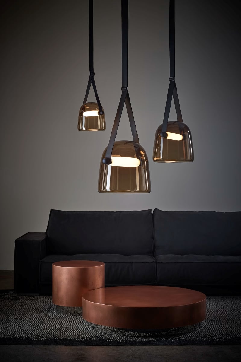 Brokis Mona pendants with glossy brown shades illuminate a dark sofa and gleaming copper tables beneath.