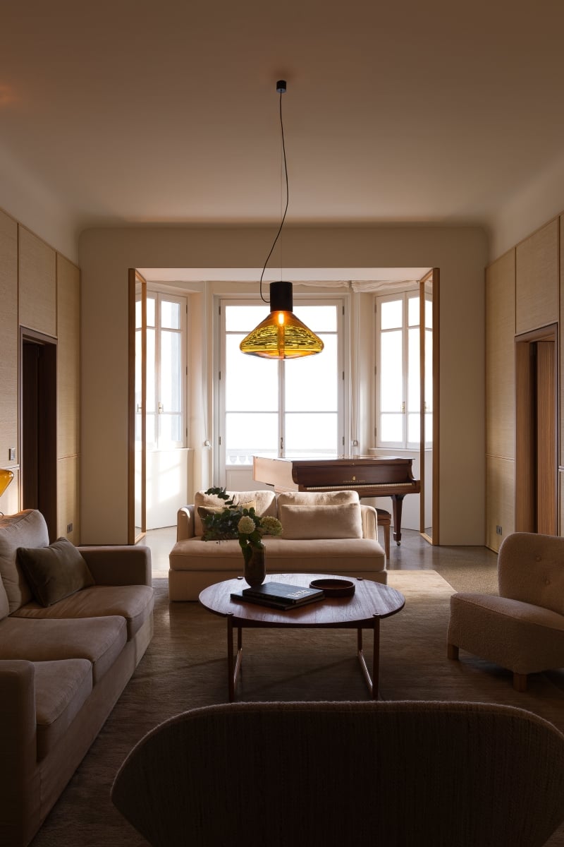 Brokis Muffins glowing over a wooden table, in a luxurious living room with beige sofas, and a grand piano.