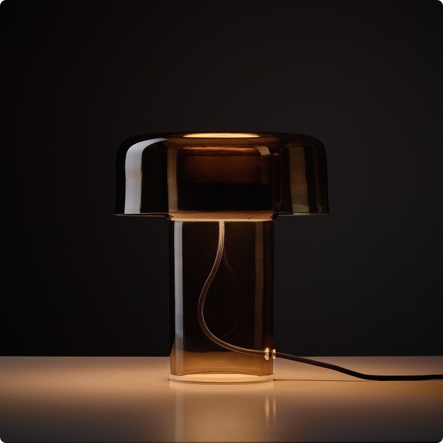 Brokis Spectra table lamp in brown handblown glass casts a soft glow, its form and flex striking on a white surface.