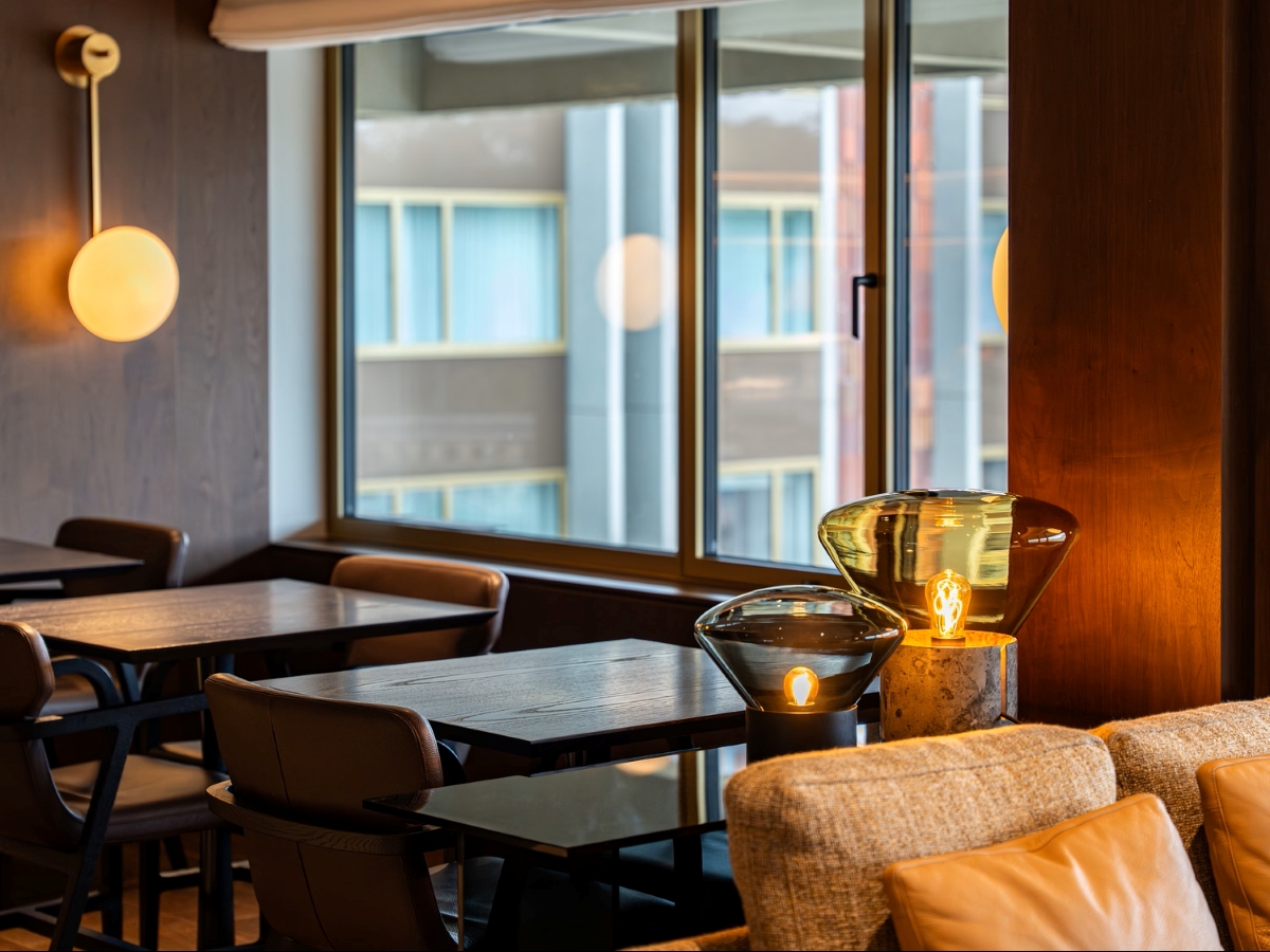 Elegant bar with dark wood tables, sofa, and Brokis Muffins lamps. Warm light, large window, city view.