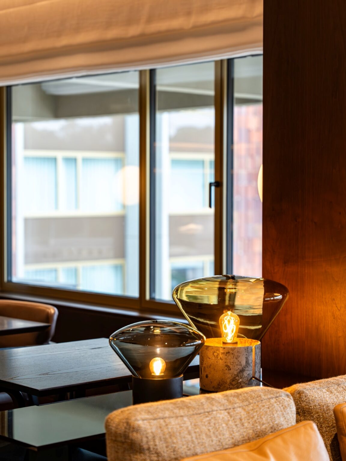 Brokis Muffins table lamps with exposed bulbs adorn a wooden table by a beige sofa, awash in sunlight from grand windows.