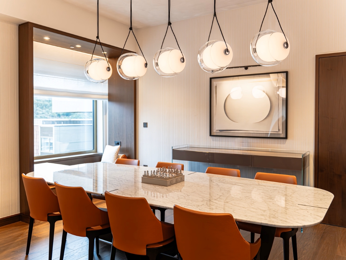 A modern dining room with a white marble table, eight orange chairs, Brokis Capsula Single pendants, and a silver chess set.