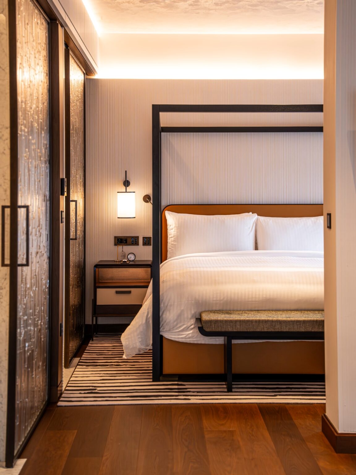 Luxurious modern hotel room with four-poster bed, crisp white bed linen, striped rug, bedside lamp, and warm lighting.