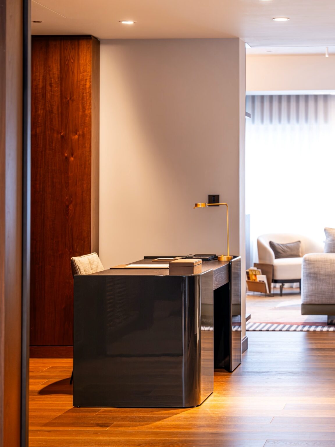 A modern home office with a glossy black desk, beige swivel chair, and sculptural gold lamp on rich wooden floors.