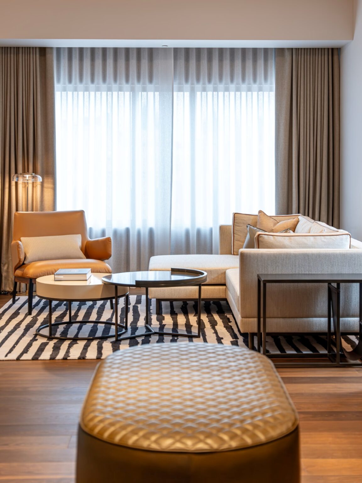 Luxurious living room with corner sofa, tan armchair, round nested tables, sheer-lit windows, and sculptural floor lamp.