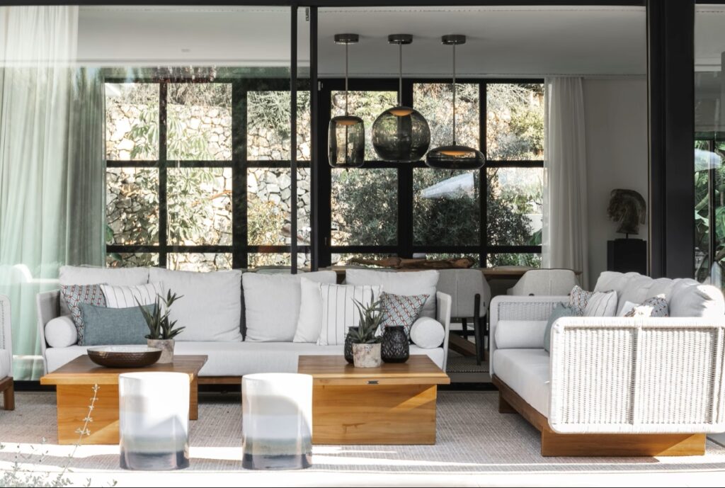 A modern patio with white-cushioned sofas, wooden accents, and lush plants opens to a dining area Brokis Knot pendants.