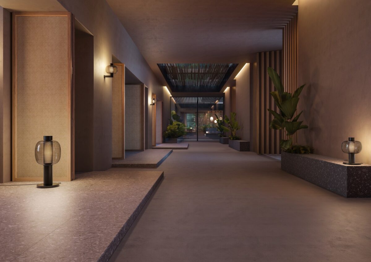 A luxe, minimalist hallway with warm light, textured beige walls, Bonbori floor lamps, and lush plants framing glass doors.