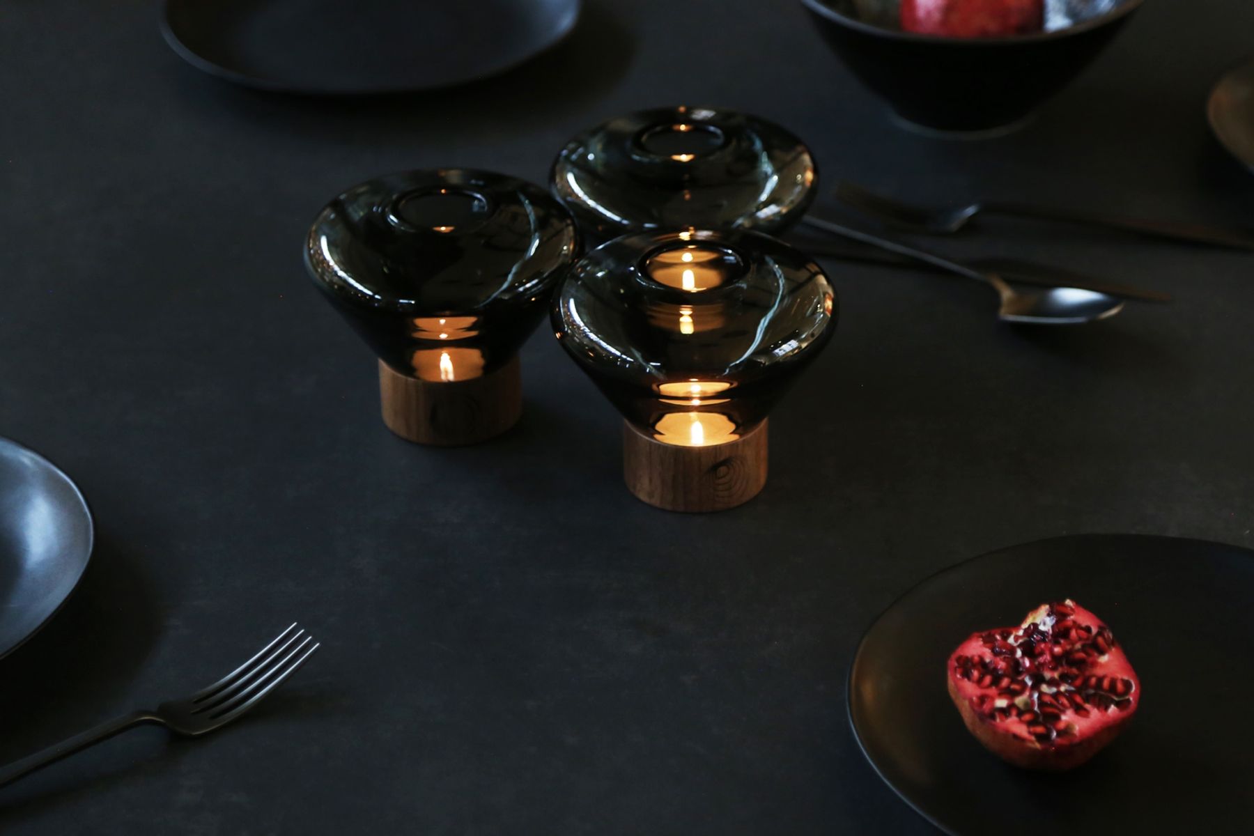 Brokis Muffins Candleholders with tea lights glow on a dark luxe table set with black plates and fresh pomegranate.