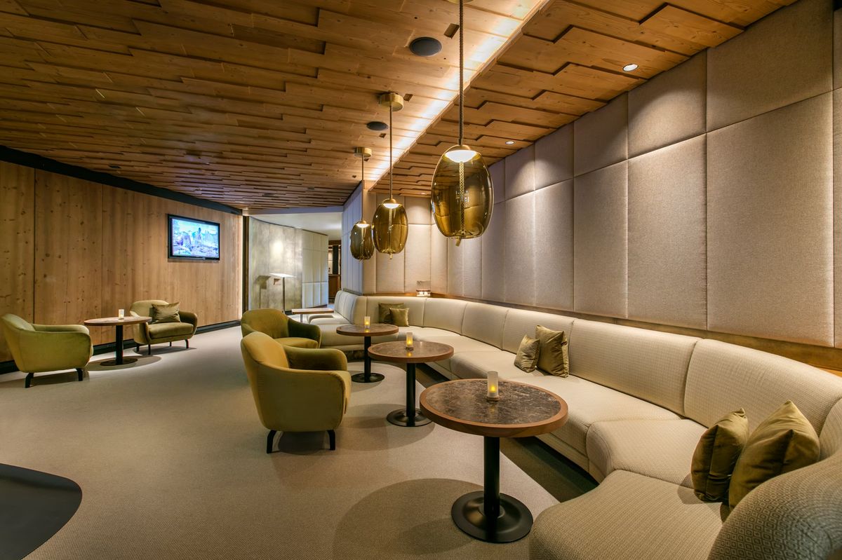 A luxe lounge with Brokis Knot pendants above round tables, green armchairs, and a long beige sofa under a wooden ceiling.