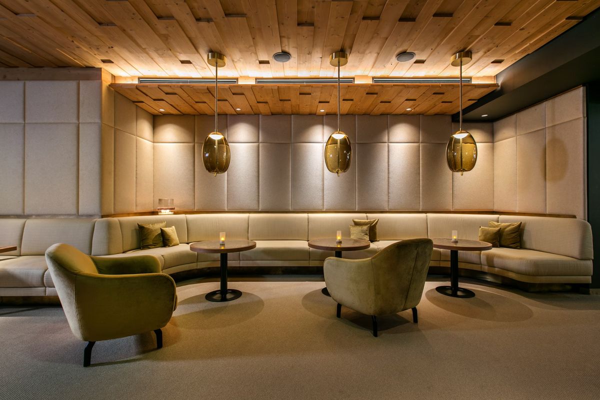 A lounge with padded walls, wooden ceiling, and Brokis Knot Uovo pendants above curved sofas and velvet chairs.