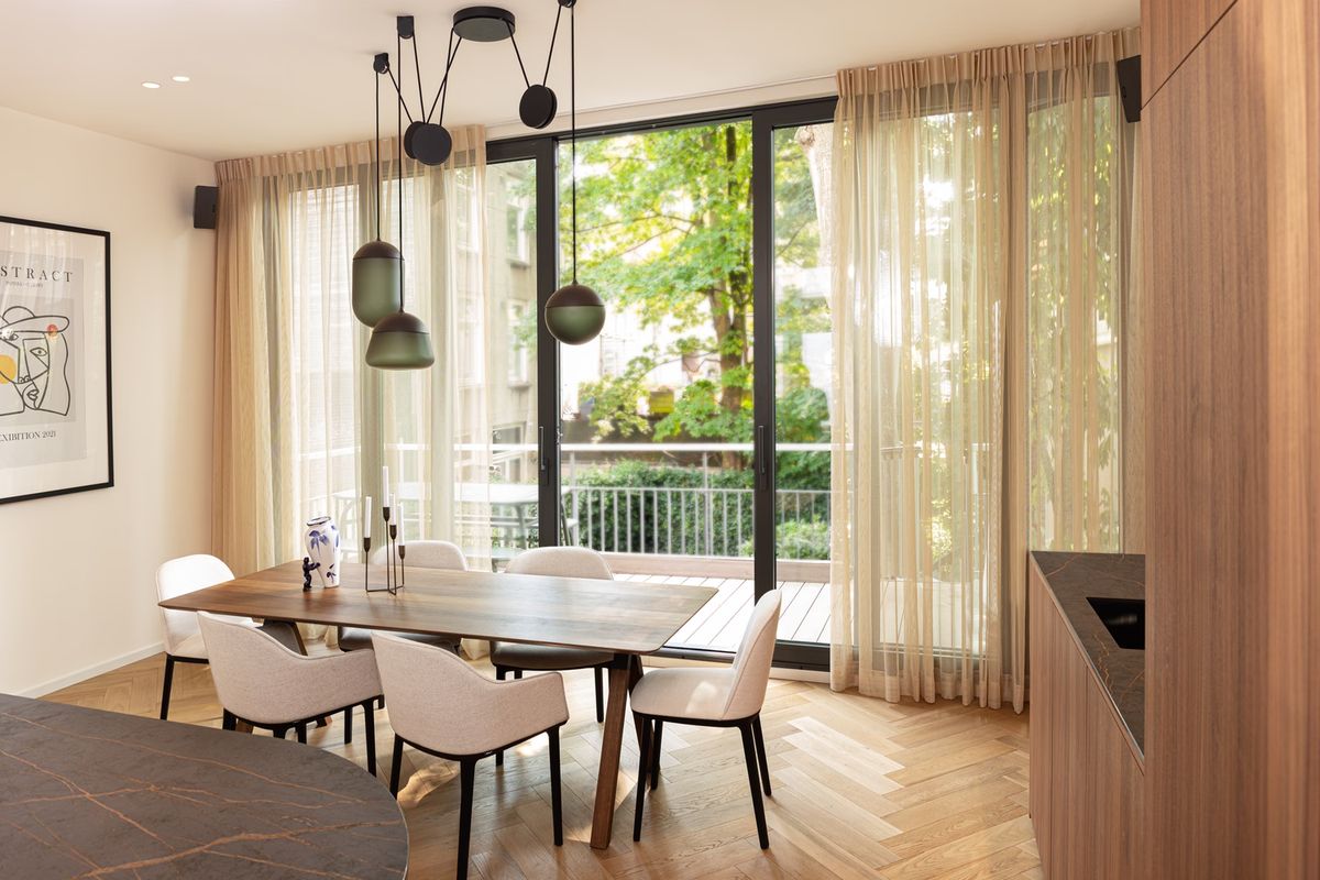 A modern dining room with a wooden table, six white chairs, Brokis Planets 3 Set, artful décor, and floor-to-ceiling windows.