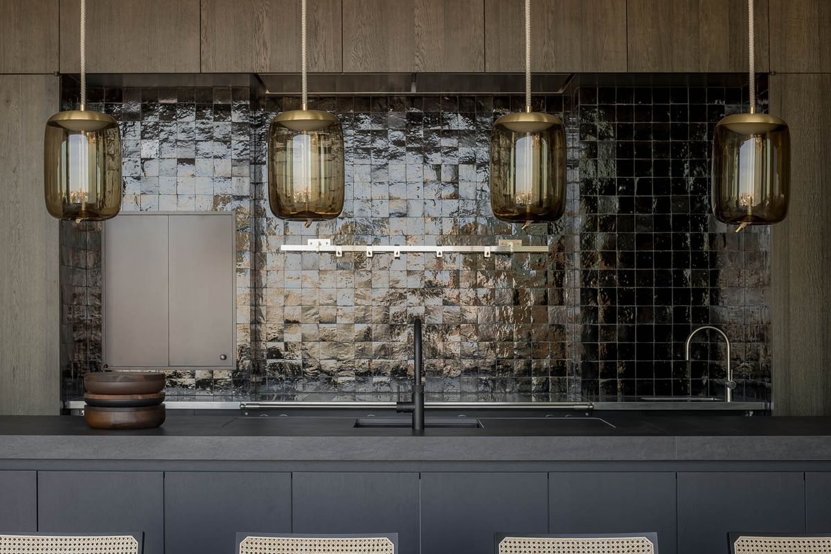 A modern kitchen with four Brokis Knot Cilindro pendants over a dark island and textured black tile splashback.