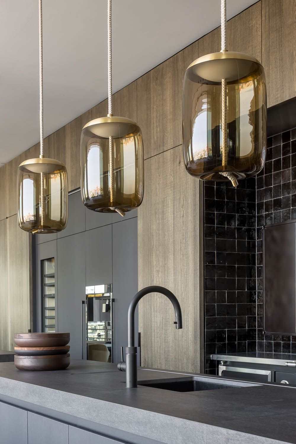 A modern kitchen with brown Brokis Knot Cilindro pendants, a sleek grey island, black tile splashback, and minimal cabinetry.