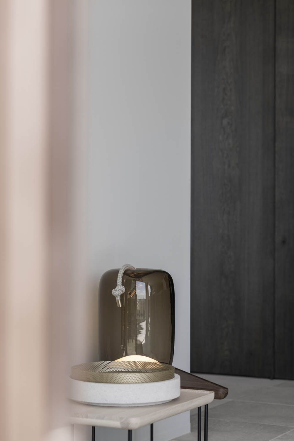 A Brokis Knot Cilindro Table with a brown glass shade graces a luxe wooden side table against contrasting modern walls.