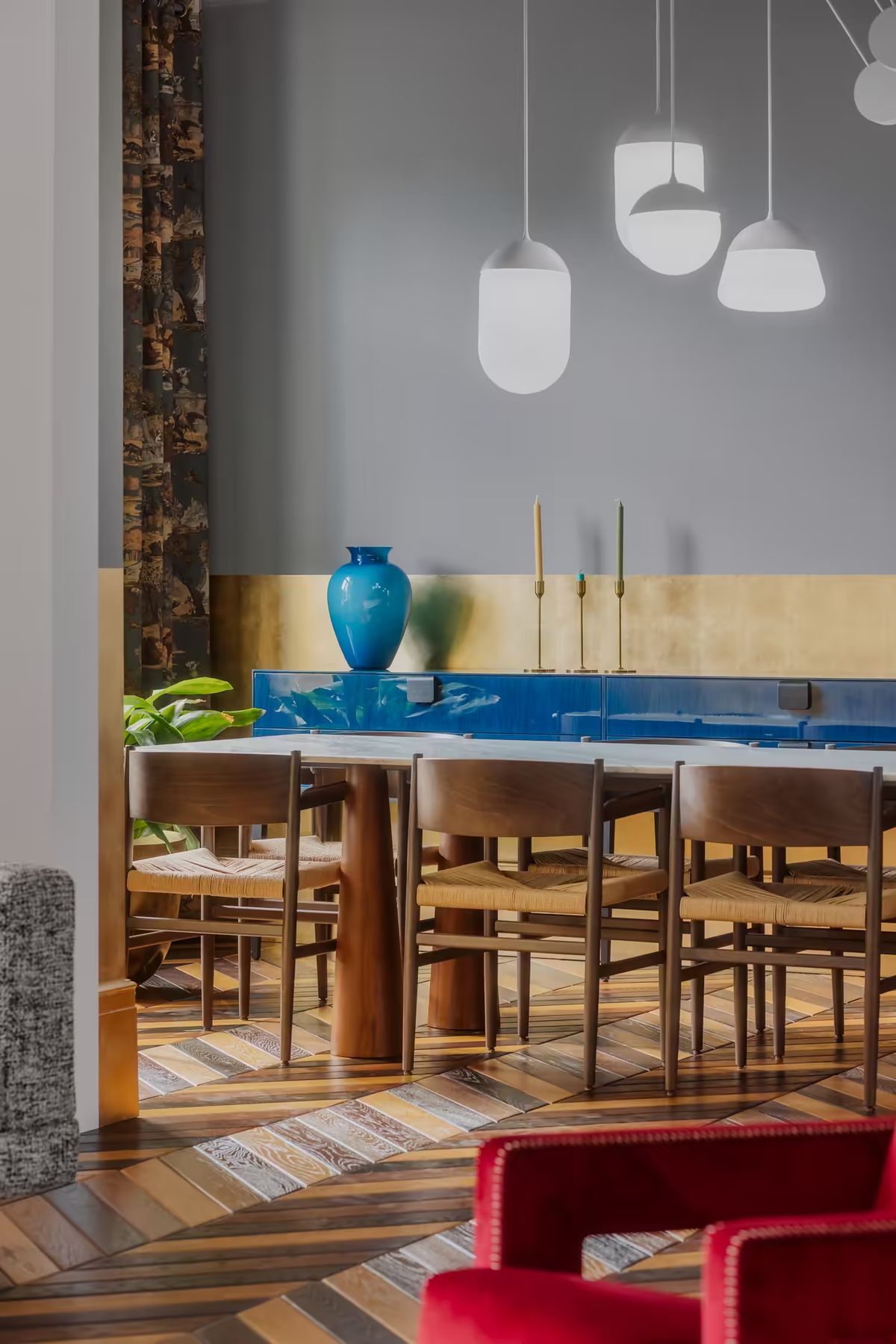 A dining room with a long wooden table, six chairs, glossy blue sideboard, gold candlesticks, and Brokis Planets 6 Set.