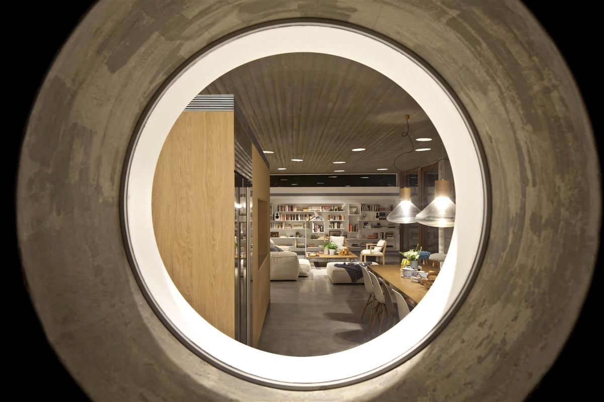Glimpse through a round concrete window into a living space with bookshelves, plush sofas, and Brokis Muffins pendants.