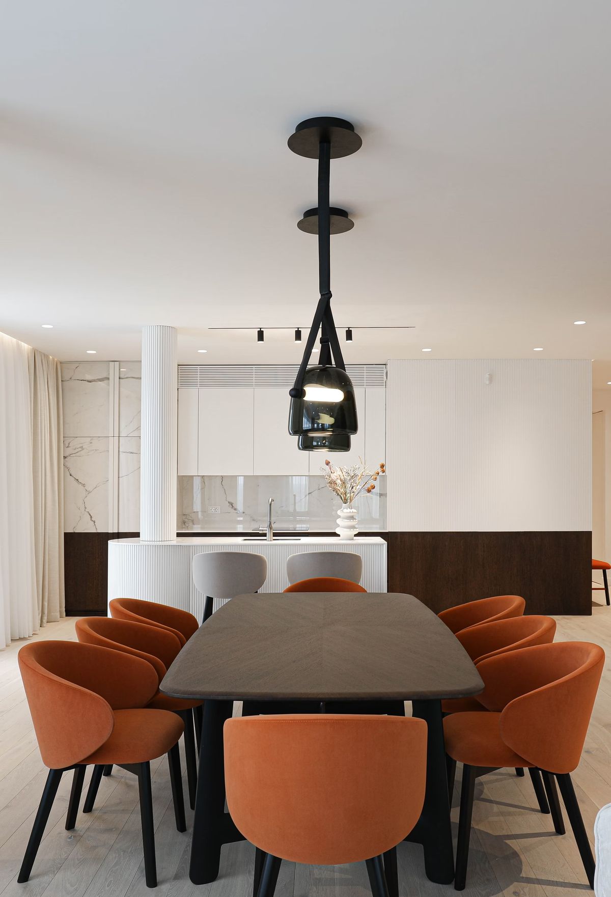 Elegant dining area with dark table, eight orange curved chairs, Brokis Mona Medium Pendants, and minimalist marble kitchen.