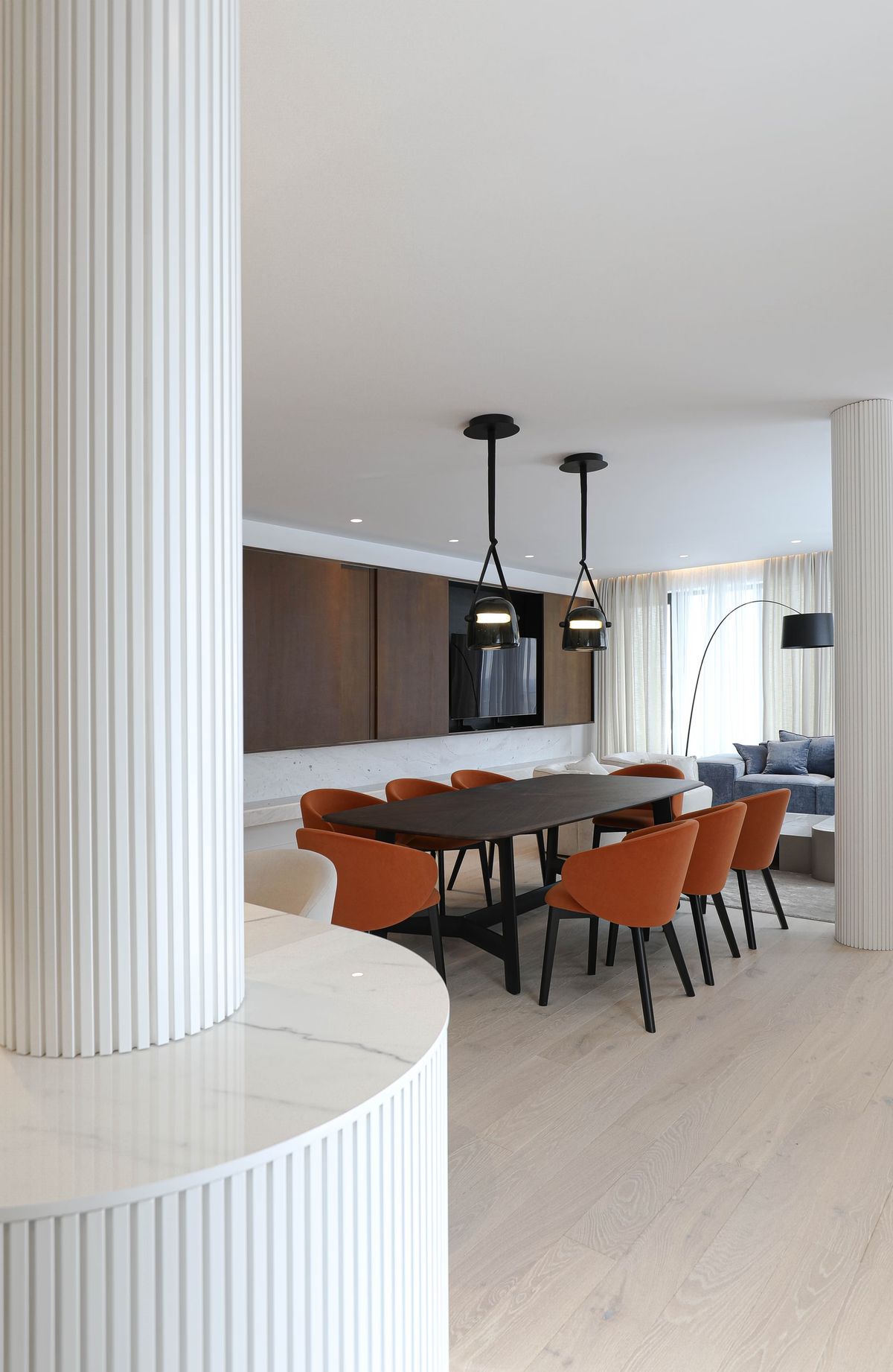 A dining room features a long dark wood table, eight rust chairs, Brokis Mona Medium Pendants, and fluted white columns.