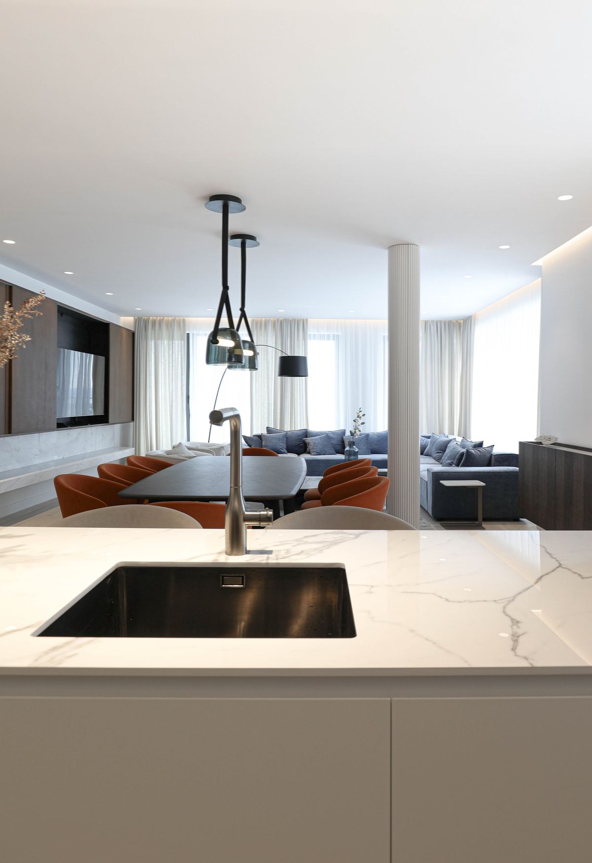 An open-plan kitchen and living room with worktops, Brokis Mona Medium Pendants, dining chairs, and sofa by large windows.