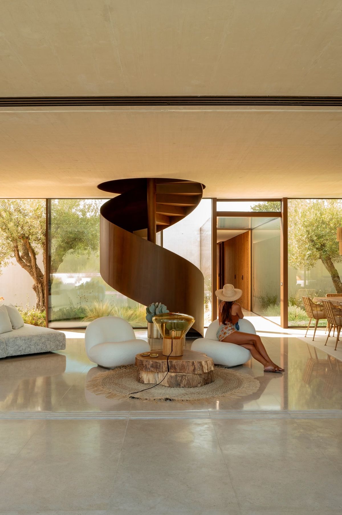 A woman in a sunhat relaxes on a chair by Brokis Muffins Wood 02; glass doors frame the tranquil garden view.