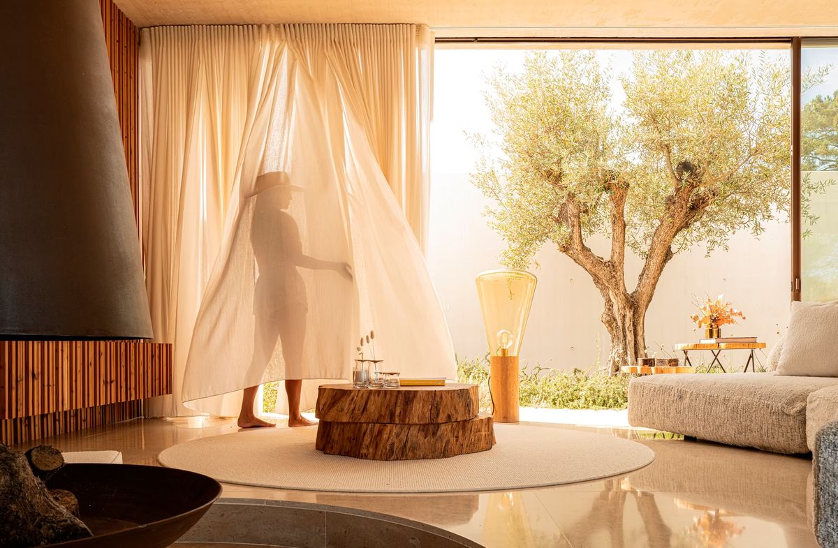 A person stands behind sheer curtains in a sunlit living room, sculpted by neutral tones and a Brokis Muffins Wood 04 Floor.