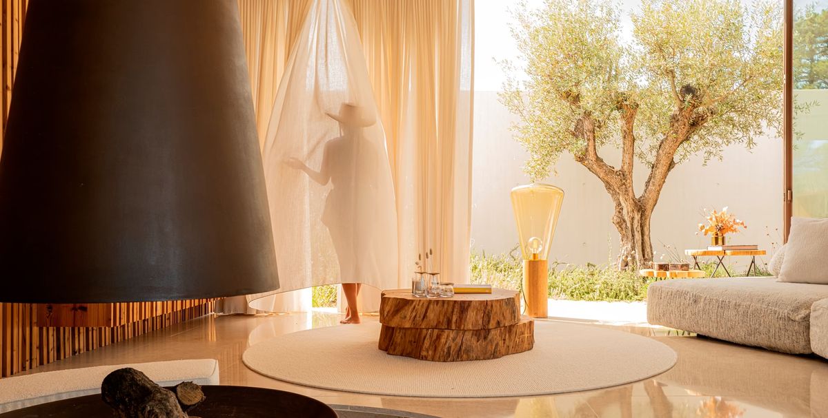 A person stands in a refined room with a Brokis Muffins Wood 04 Floor, a graceful tree, and elegant draped curtains.