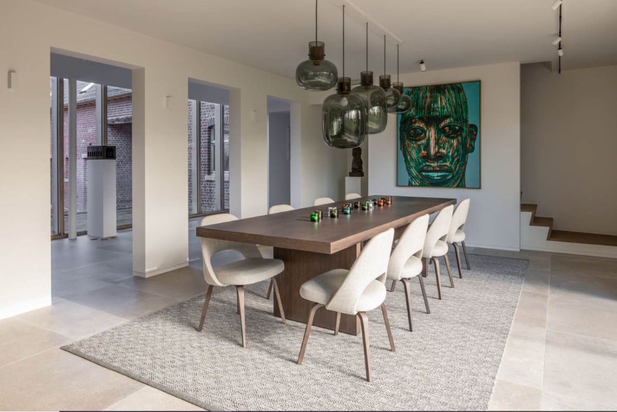 A modern dining room with a long wooden table, ten pale chairs, textured rug, Brokis Awa pendants, and green portrait.