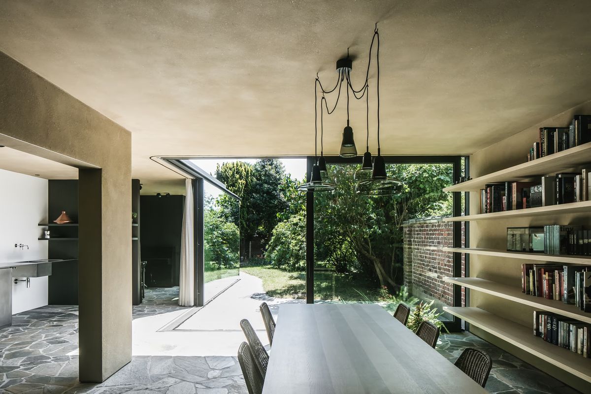A chic dining room with a long table, woven chairs, Brokis Shadows Set Free, stone floor, and glass doors to a lush garden.